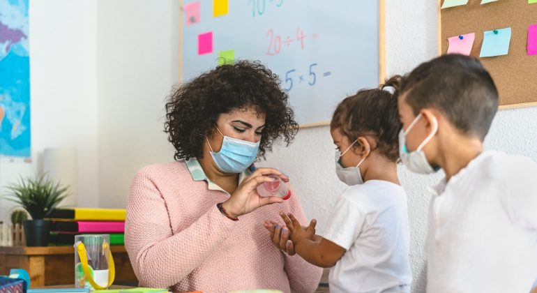Teacher cleaning hands to student children with sanitizer gel while wearing face mask